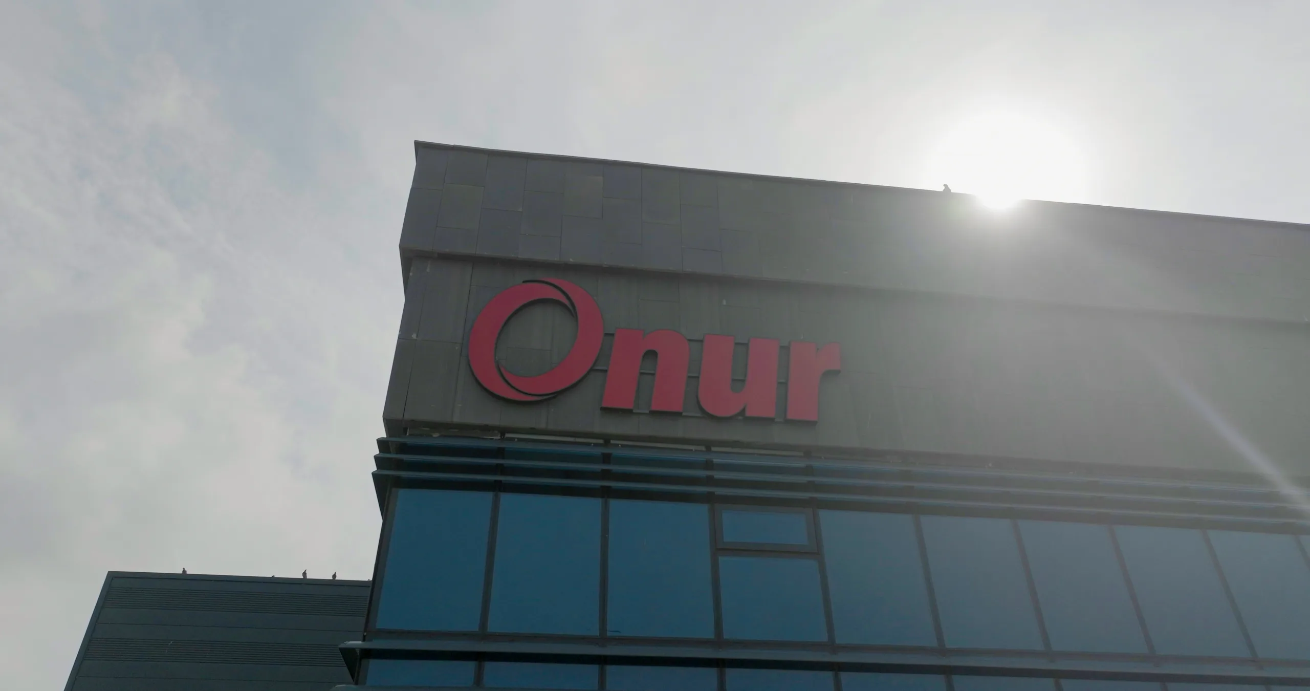 Yakın plan: koyu renkli bina cephesinde kırmızı "Onur" tabelası; alttan, güneşe karşı çekilmiş.
