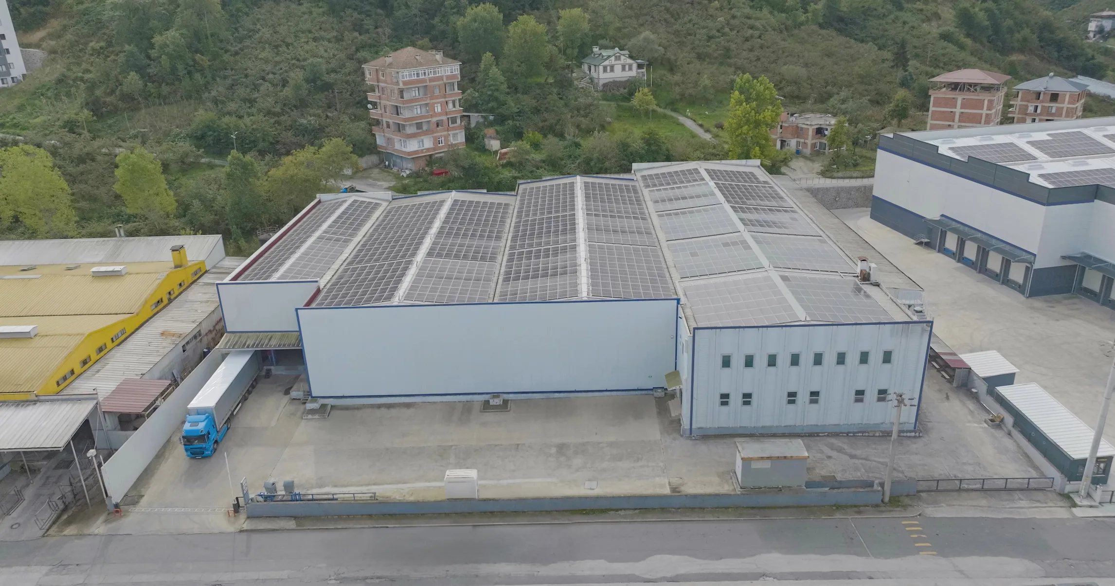 Havadan görünüm: güneş panelli çatısıyla uzun bir depo binası, yanında yükleme alanı ve yola paralel uzanan servis yolu.